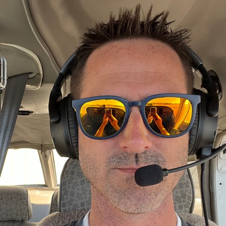 Paul Brown in the cockpit of a small airplane, wearing an aviation headset and mirrored sunglasses